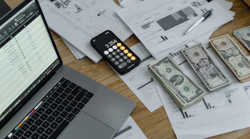 A desk with financial documents, currency, a laptop, and phone calculator.
